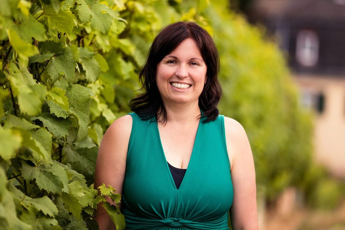 Frau steht lächelnd neben einer Weinrebe oder Hecke