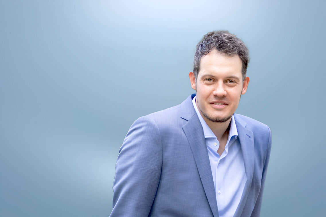 Mann in Anzug steht entspannt auf etwas gelehnt da und schaut lächelnd in die Kamera vor verschwommenem Hintergrund