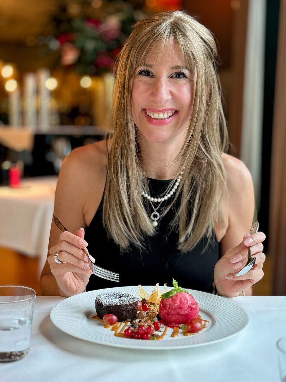 Kobieta siedząca przy stole i trzymająca w dłoniach łyżkę i widelec, śmiejąca się do kamery, przed nią talerz z deserami.