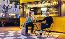 Frau, Mann und Hund sitzen vor einem Kiosk und prosten sich zu.
