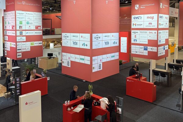 Messestand in rot neben weiteren Ständen im Hintergrund, darüber jeweils eine Infotafeln mit abgebildeten Firmenlogos, an den Ständen stehen einige Personen