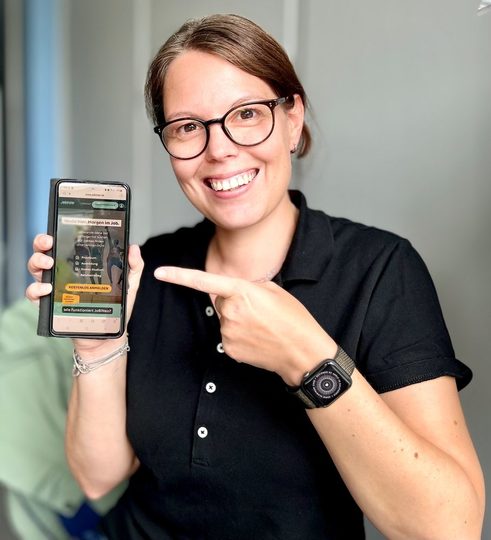 Frau mit Brille zeigt lächelnd auf Handy