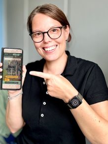 Frau mit Brille zeigt lächelnd auf Handy
