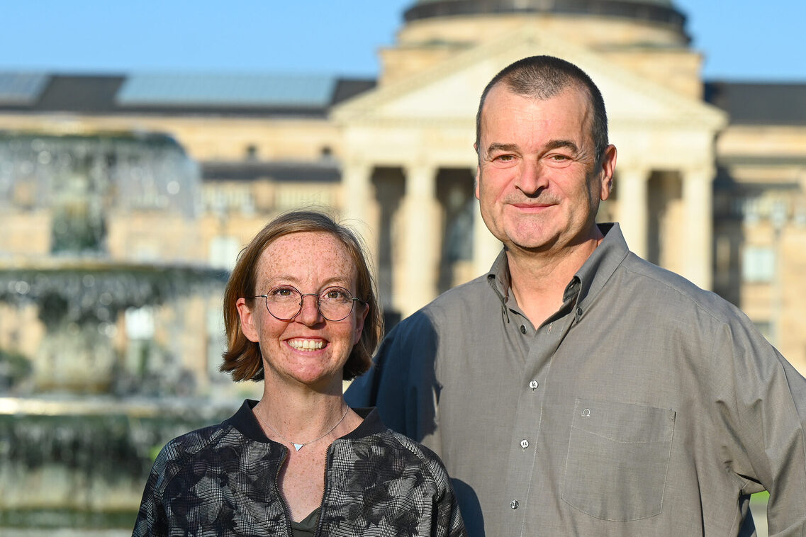 eine Frau und ein Mann stehen vor einem Brunnen und antik wirkendendem Gebäude im verschwommenen Hintergrund