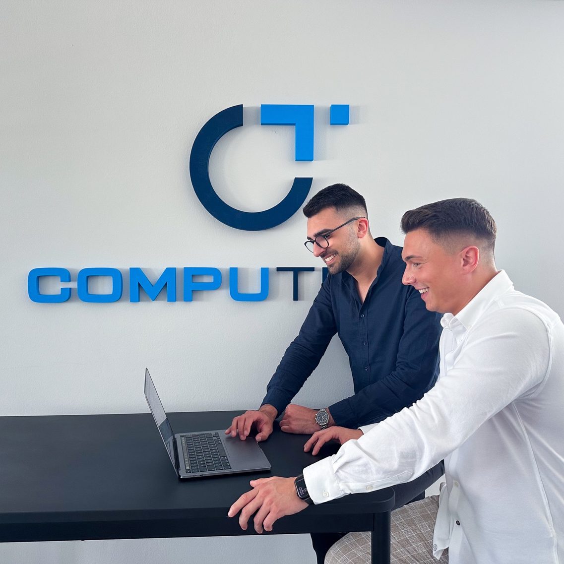Two men are standing at a table, looking at an open laptop together. In the background, a company logo is visible on a white wall.