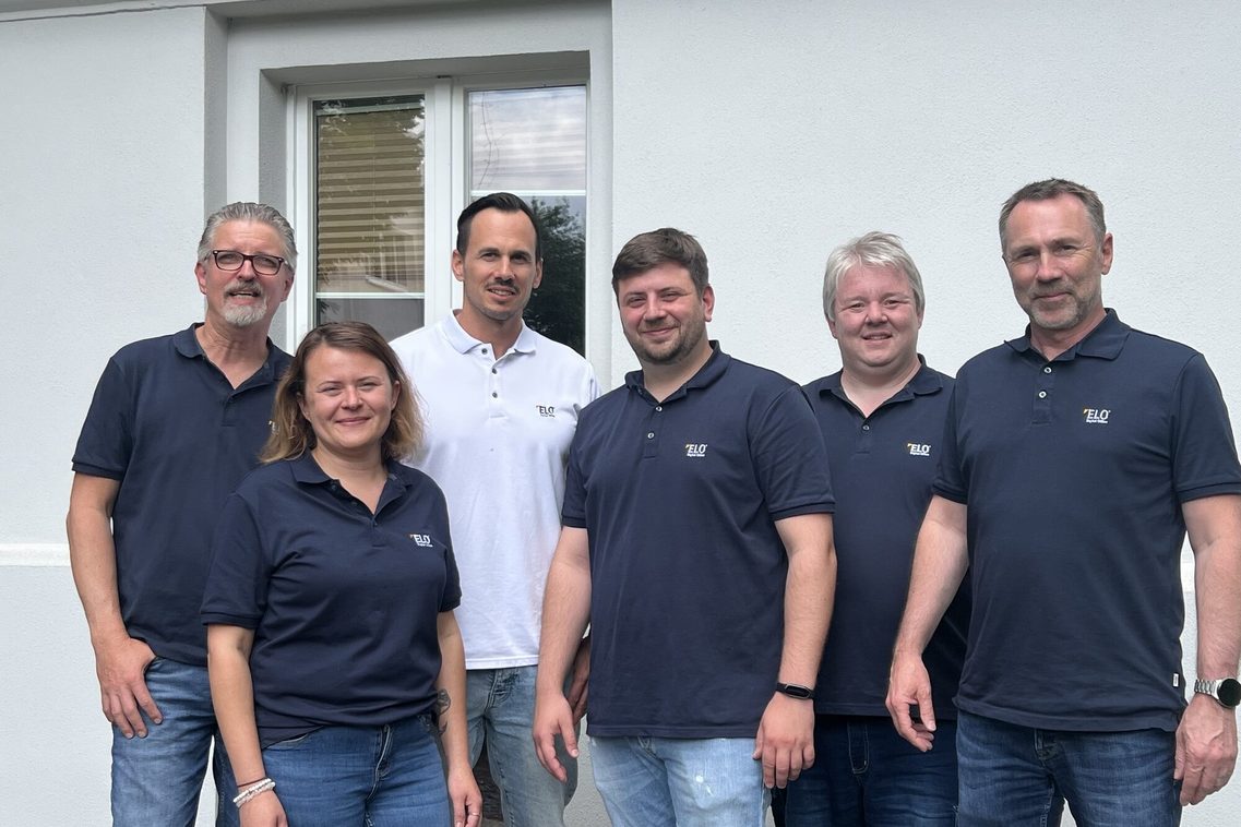 Sechs Personen stehen vor einer hellen Hauswand mit einem Fenster im Hintergrund und blicken in die Kamera.