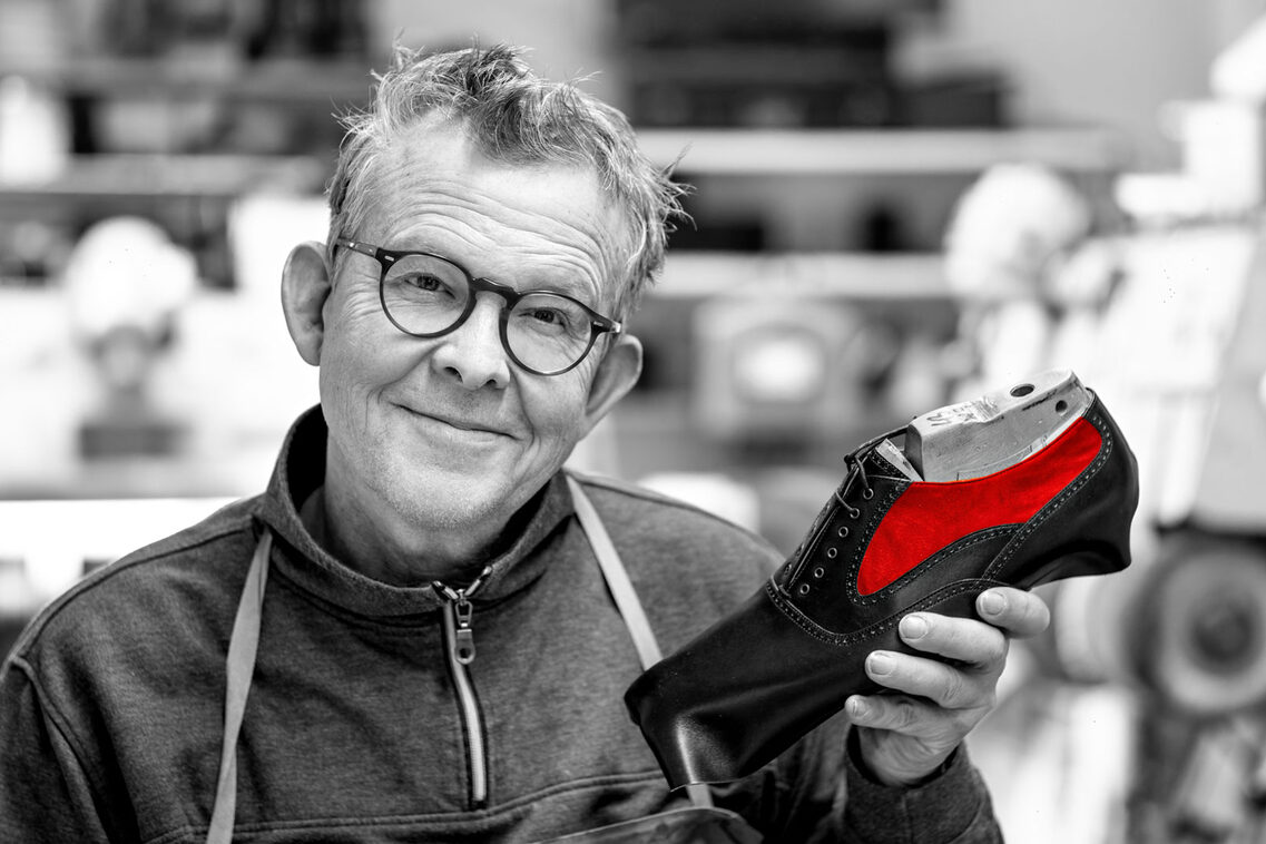 Mann hält in der Hand einen Lederschuh mit rotem Akzent, bis auf den Akzent ist das Bild in Schwarz-Weiß