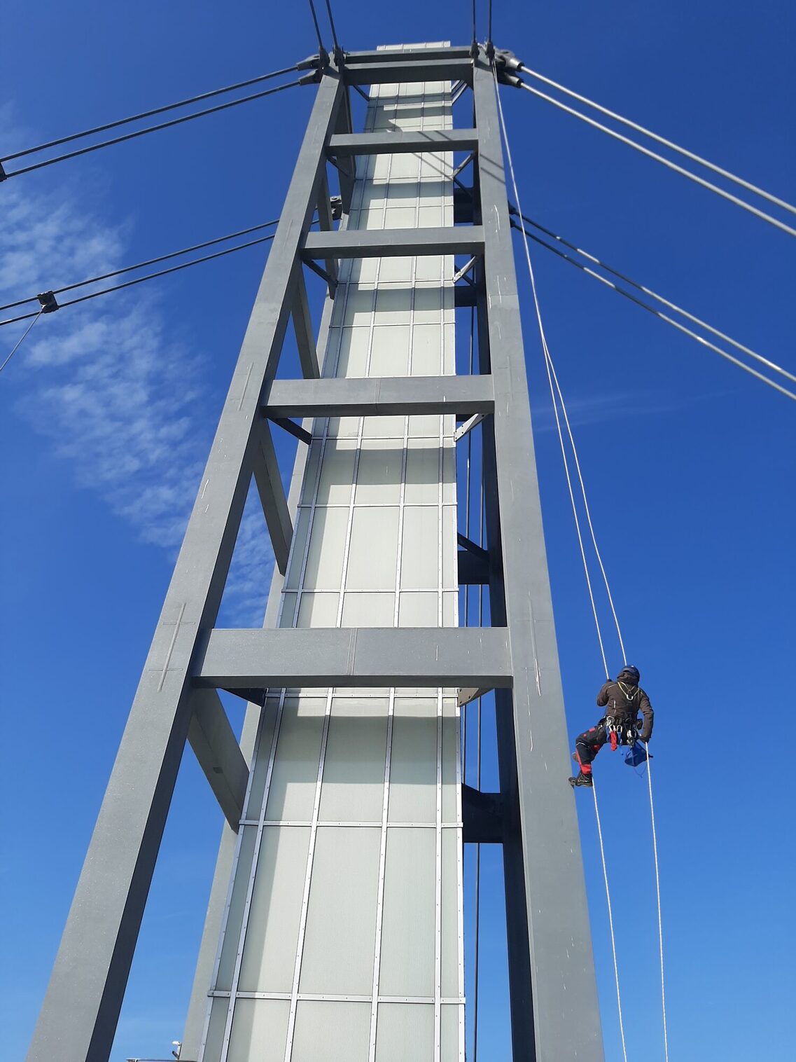 ein Mensch hängt an Seilzug an einem Metallrahmen eines turmähnlichen Gebildes, von dessen Spitze gehen Stahlseile ab