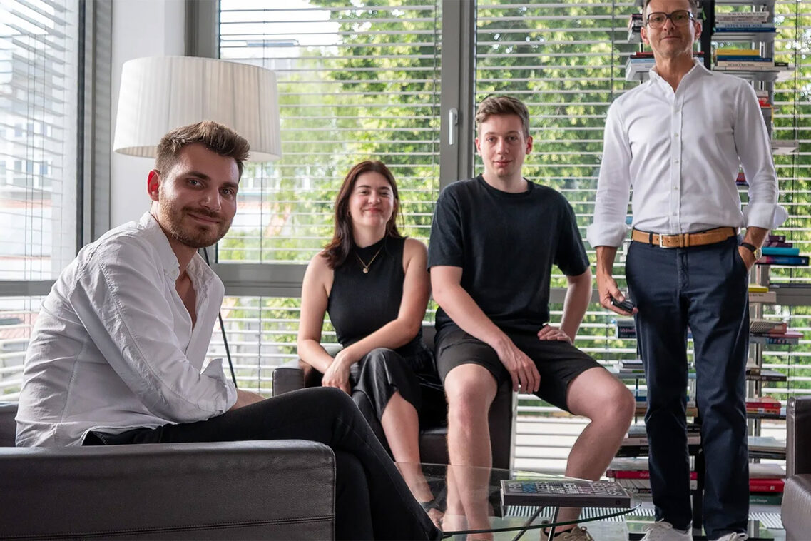 Eine Frau und drei Männer auf Sesseln und im Stehen.