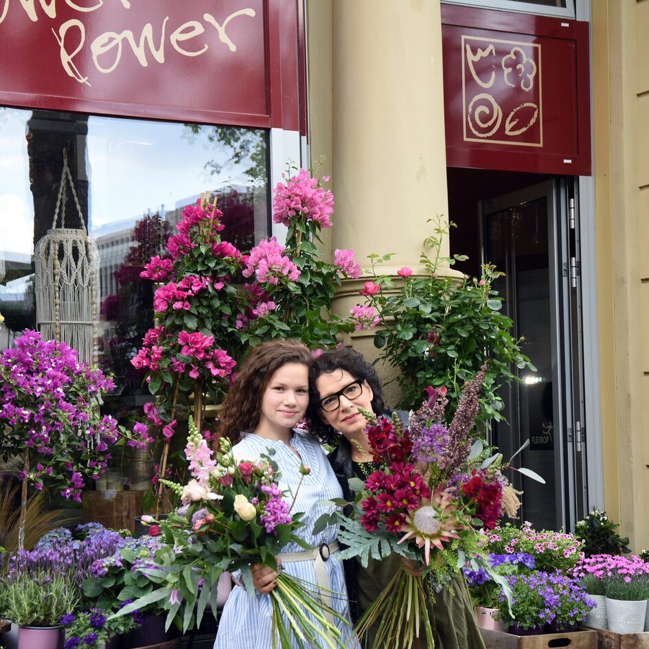zwei Frauen stehen Kopf an Kopf vor einem Blumenladen mit der Fensteraufschrift "FlowerPower" und einigen Sträußen und Töpfen mit blühenden Pflanzen und halten Blumensträuße in der Hand