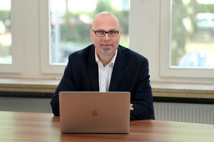 Mann in Anzug und Brille sitzt lächelnd hinter offenem Laptop an Tischfläche mit Holzmaserung vor einer hellen Fensterfront