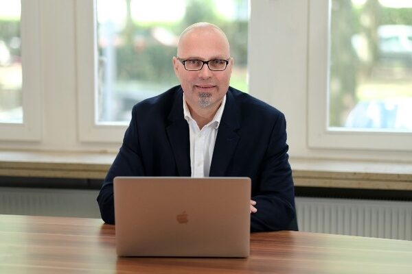 Un uomo in giacca e cravatta e occhiali siede sorridente dietro un computer portatile aperto a un tavolo con venature di legno davanti a una finestra luminosa